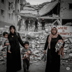 Grayscale image showing a family, including a mother holding a child and another child with a toy, standing amidst the extensive rubble of destroyed buildings in a conflict zone, with a sign in the background asking "WHERE IS HUMANITY?". Rescue workers are visible searching through debris in the distance.