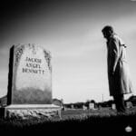 Noir black and white of Jack Hammer standing by the headstone of Jackie Angel Bennett