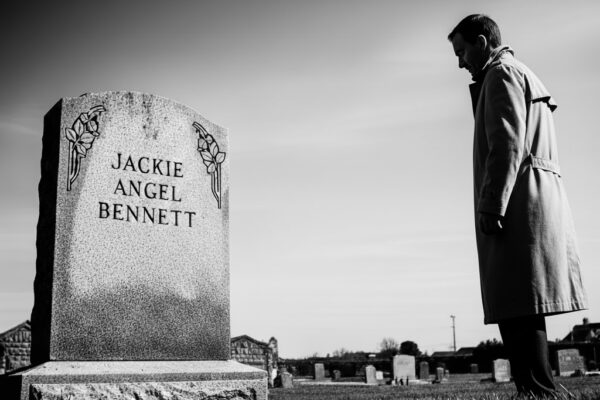 Noir black and white of Jack Hammer standing by the headstone of Jackie Angel Bennett