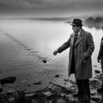 Noir. Black and white. A noir detective and his 20 year old, Punk Rock daughter stand in front of a country lake that stretches to the horizon. The detective has skipped a stone across the lake and the stone goes on forever.