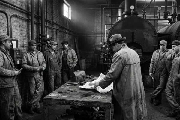 Noir. Black and white. Factory men with timecards standing along the wall of a boiler room in groups of three and four near a time clock as a detective spreads open a newspaper on a rusted steel table.